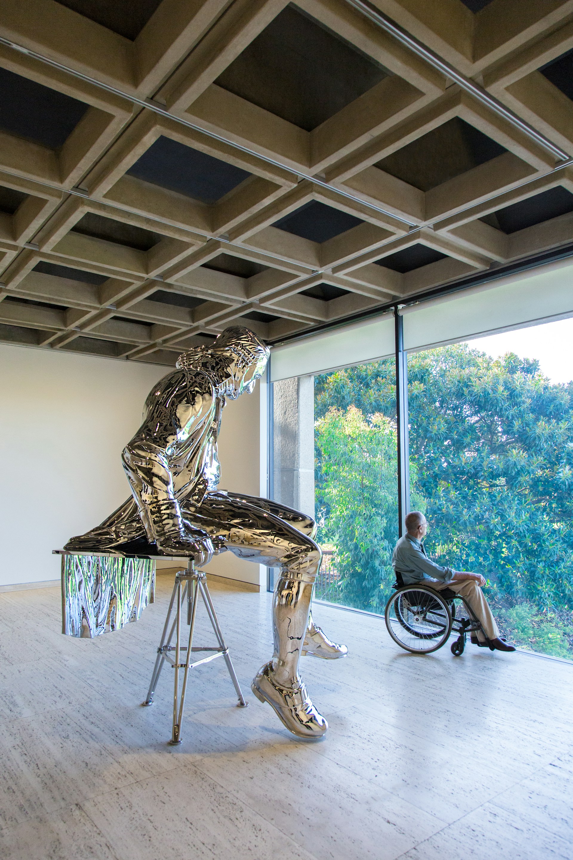Spiegelende kunstsculptuur in museumruimte met op de achtergrond een persoon in een rolstoel bij een groot raam